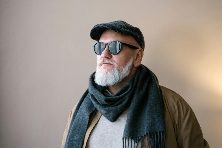 Portrait of a stylish elderly man wearing sunglasses, hat, and scarf in an indoor setting.